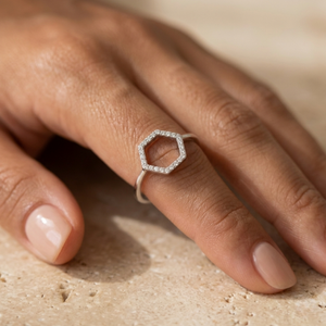 A silver hexagonal ring with small clear stones, worn on a finger. The hand is resting on a textured beige stone surface.